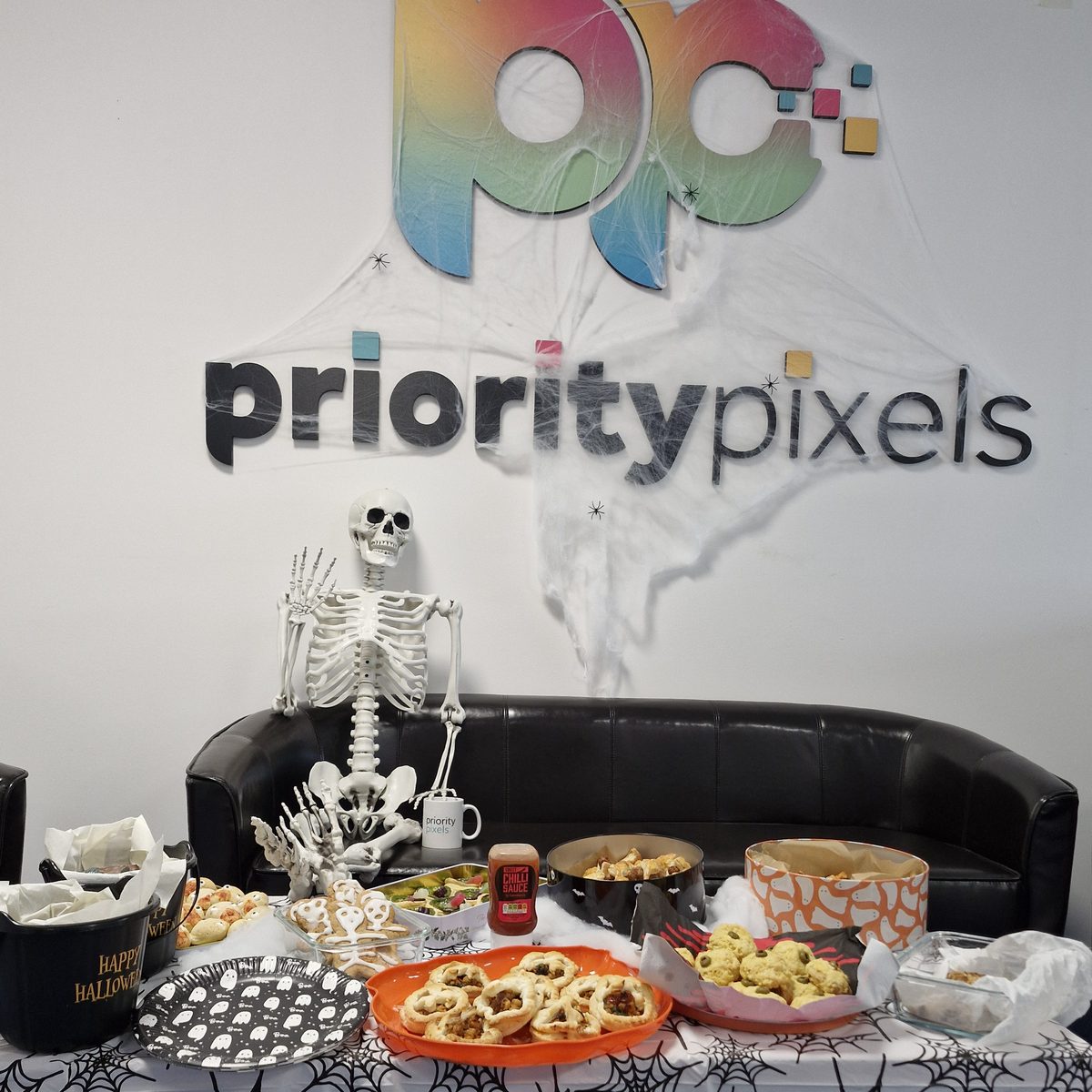 Halloween bake table in front of the Priority Pixels logo wall covered in cobwebs, with a skeleton on the sofa
